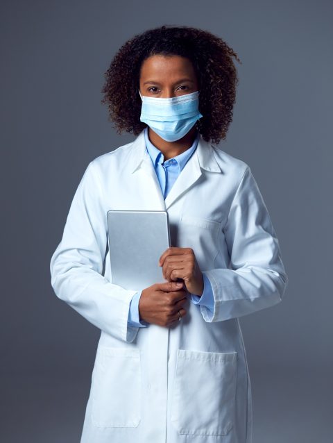 Studio Portrait Of Female Doctor In Lab Coat Wearing Face Mask Holding Digital Tablet